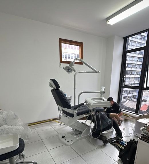 Cadeira odontológica sendo instalada por dois técnicos numa sala branca com janela.